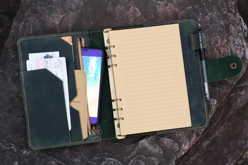 May include: A dark green leather bound journal with a zippered pocket, a phone, and a pen. The journal has a lined paper insert and a ring binder.
