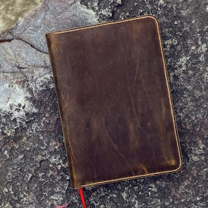 May include: A brown leather notebook with a gold stitched border. The notebook is lying on a rough, gray rock surface.