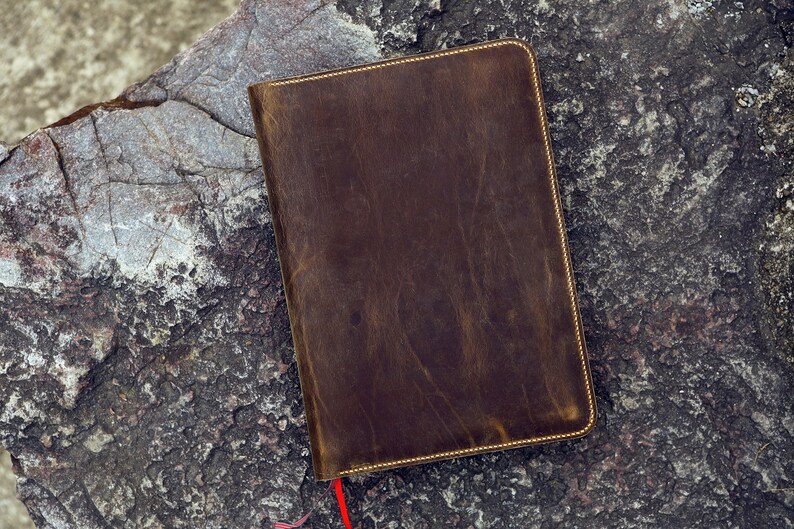 May include: A brown leather notebook with a gold stitched border. The notebook is lying on a rough, gray rock surface.