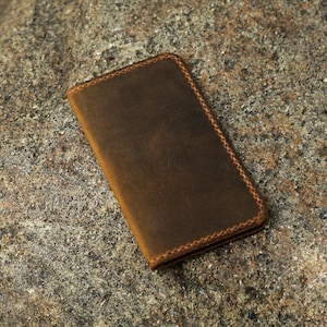 May include: A brown leather wallet with a stitched edge. The wallet is lying on a rough, textured surface.