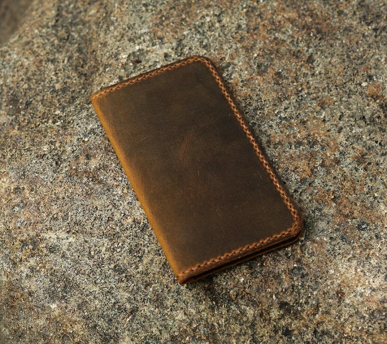 May include: A brown leather wallet with a stitched edge. The wallet is lying on a rough, textured surface.