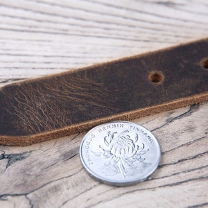 May include: Close-up of a brown leather belt with two holes. The belt is lying on a light wood surface. A silver Chinese coin with the text "Zhongguo Renmin YinHang" is in the foreground.