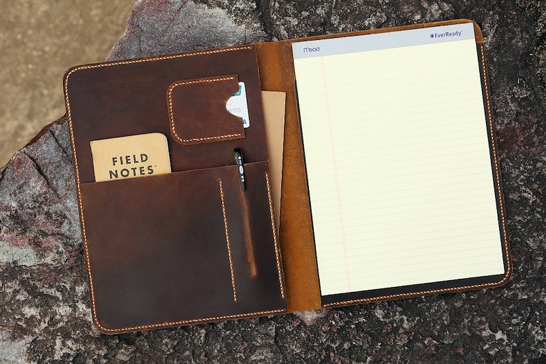 May include: A brown leather portfolio with a pen holder, a pocket for a notepad, and a lined notepad that says "EverReady" on the top right corner. The notepad also has the word "Mead" printed on the top left corner.