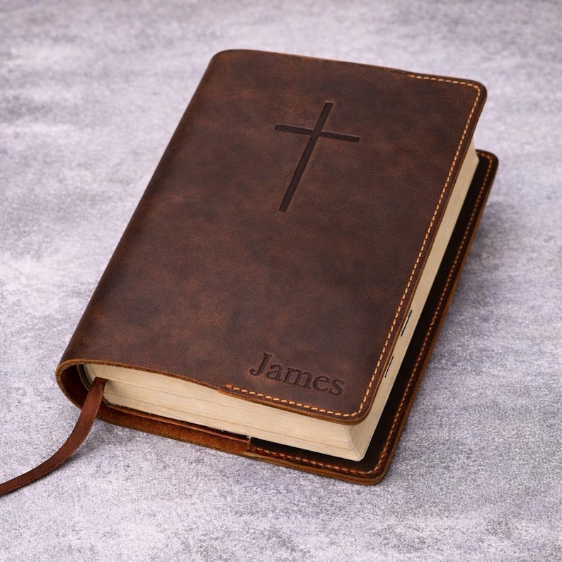 May include: A brown leather-bound book with a cross embossed on the cover. The word "James" is imprinted at the bottom. The book has a sewn binding and a brown ribbon bookmark.