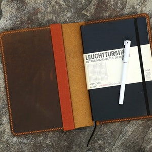 Puede incluir: Una cubierta de cuaderno de cuero marr&oacute;n con una solapa de cuero marr&oacute;n claro y naranja. La cubierta est&aacute; cerrada y un cuaderno negro con un bol&iacute;grafo blanco est&aacute; dentro. El cuaderno tiene el texto "LEUCHTTURM1917" y "DETAILS MAKE ALL THE DIFFERENCE" en la parte delantera.