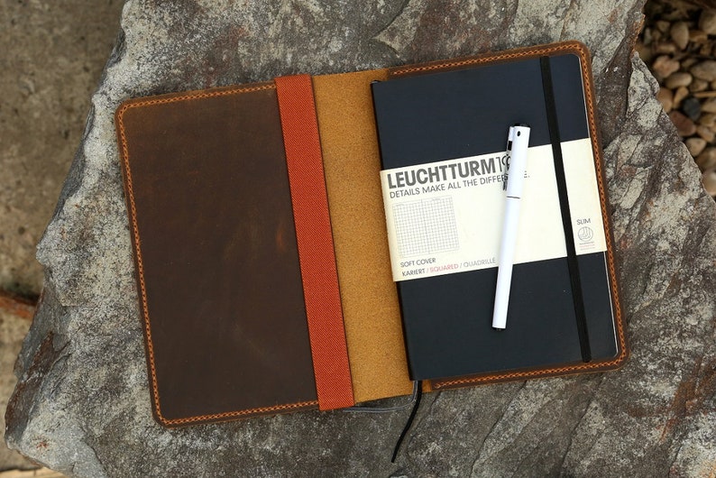Puede incluir: Una cubierta de cuaderno de cuero marr&oacute;n con una solapa de cuero marr&oacute;n claro y naranja. La cubierta est&aacute; cerrada y un cuaderno negro con un bol&iacute;grafo blanco est&aacute; dentro. El cuaderno tiene el texto "LEUCHTTURM1917" y "DETAILS MAKE ALL THE DIFFERENCE" en la parte delantera.