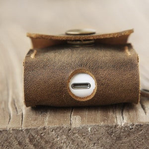 May include: Close-up of a brown leather case with a button closure. The case has a distressed texture and a circular opening with a white interior. The case is designed to hold a small electronic device.