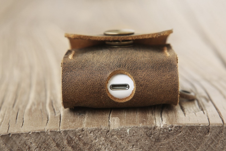 May include: Close-up of a brown leather case with a button closure. The case has a distressed texture and a circular opening with a white interior. The case is designed to hold a small electronic device.