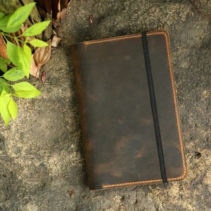 May include: A brown leather notebook with a black elastic closure. The notebook has a distressed look and is stitched with orange thread.