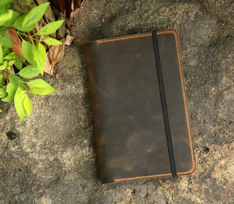 Puede incluir: Un cuaderno de cuero marr&oacute;n con cierre el&aacute;stico negro. El cuaderno tiene un aspecto desgastado y est&aacute; cosido con hilo naranja.