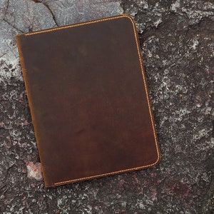 May include: A brown leather notebook with a stitched edge. The notebook is lying on a rough, textured surface.