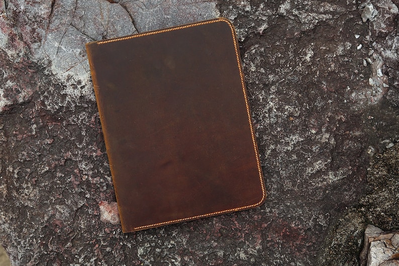 May include: A brown leather notebook with a stitched edge. The notebook is lying on a rough, textured surface.