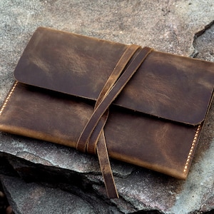 Könnte beinhalten: Eine braune Ledertasche mit einem Bindeverschluss. Die Tasche ist abgenutzt und hat einen rustikalen Look.