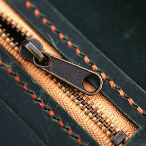 May include: Close-up of a zipper on a dark green leather bag. The zipper is brass colored and has the text "YKK" on it. The bag is stitched with orange thread.
