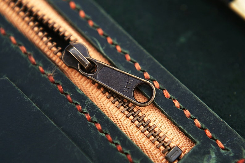 May include: Close-up of a zipper on a dark green leather bag. The zipper is brass colored and has the text "YKK" on it. The bag is stitched with orange thread.