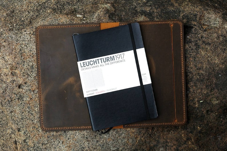 Puede incluir: Una cubierta de cuaderno de cuero marr&oacute;n con un cuaderno negro en el interior. El cuaderno tiene el texto "LEUCHTTURM1917" y "DETAILS MAKE ALL THE DIFFERENCE" en la portada.