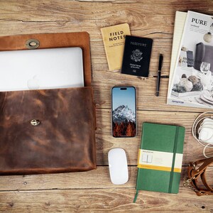 May include: A brown leather laptop bag with a flap closure and a metal buckle. The bag is open and a laptop computer is inside. Other items on the table include a passport, a notebook, a phone, a mouse, and a pen.