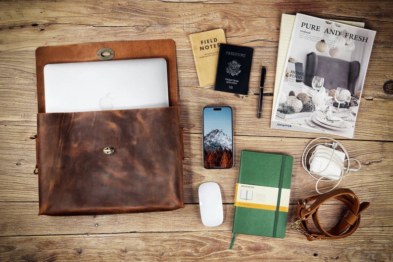 May include: A brown leather laptop bag with a flap closure and a metal buckle. The bag is open and a laptop computer is inside. Other items on the table include a passport, a notebook, a phone, a mouse, and a pen.