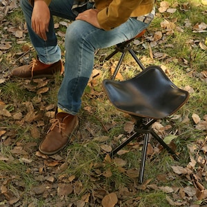 Puede incluir: Un taburete de camping de cuero negro con base de tr&iacute;pode. El taburete est&aacute; hecho de materiales duraderos y es perfecto para actividades al aire libre.