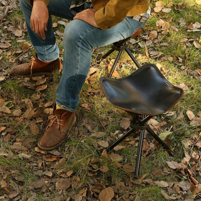Puede incluir: Un taburete de camping de cuero negro con base de tr&iacute;pode. El taburete est&aacute; hecho de materiales duraderos y es perfecto para actividades al aire libre.