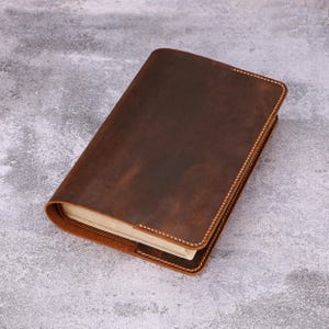 May include: A brown leather journal with a stitched border. The journal is open, revealing the pages inside. The leather has a distressed look, and the stitching is a light tan color. The journal is rectangular and appears to be a notebook or diary.