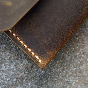 Op de afbeelding: Close-up van een bruine leren portemonnee met witte stiksels. De portemonnee is gemaakt van verweerd leer en heeft een rustieke uitstraling.