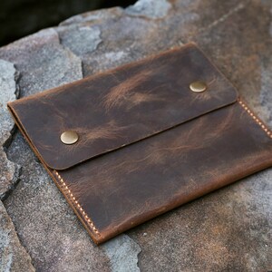 Puede incluir: Una cartera de cuero marr&oacute;n con dos broches de presi&oacute;n de lat&oacute;n y un borde cosido.