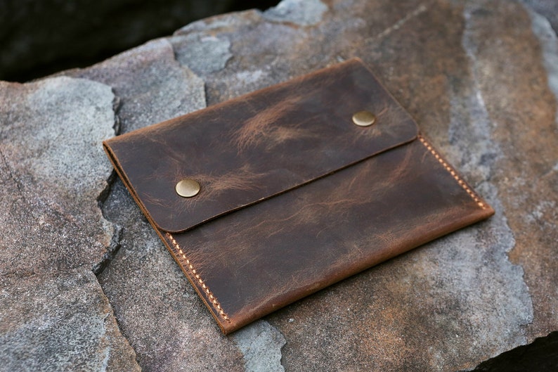 Puede incluir: Una cartera de cuero marr&oacute;n con dos broches de presi&oacute;n de lat&oacute;n y un borde cosido.