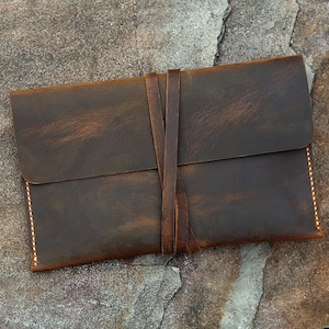 May include: A brown leather envelope with a flap closure and a tied leather strap. The envelope has a distressed look and is stitched with a light brown thread.