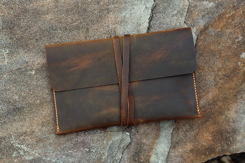 May include: A brown leather envelope with a flap closure and a tied leather strap. The envelope has a distressed look and is stitched with a light brown thread.