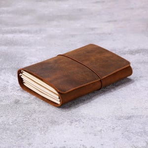 Op de afbeelding: Een bruin leren notitieboek met een riemsluiting. Het notitieboek is rechthoekig en heeft een versleten, getextureerde uitstraling. De pagina's zijn zichtbaar en lijken leeg te zijn. Het notitieboek ligt op een grijze ondergrond.