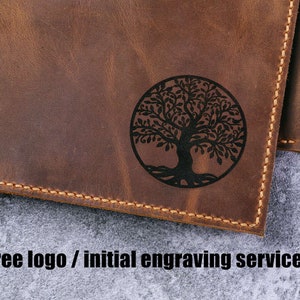 May include: A brown leather wallet featuring a circular black engraved tree of life design on the front surface. The wallet has visible tan stitching along the edges. The text "Free logo / initial engraving service" is printed below the wallet.