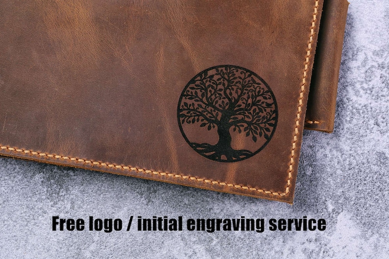 May include: A brown leather wallet featuring a circular black engraved tree of life design on the front surface. The wallet has visible tan stitching along the edges. The text "Free logo / initial engraving service" is printed below the wallet.