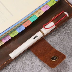 May include: A brown leather notebook cover with a snap closure and a pen loop holding a white fountain pen with a red wire clip. The notebook features colorful monthly index tabs labeled Feb, Mar, Apr, May, Jun, Jul, Aug, and Sep along the edge.