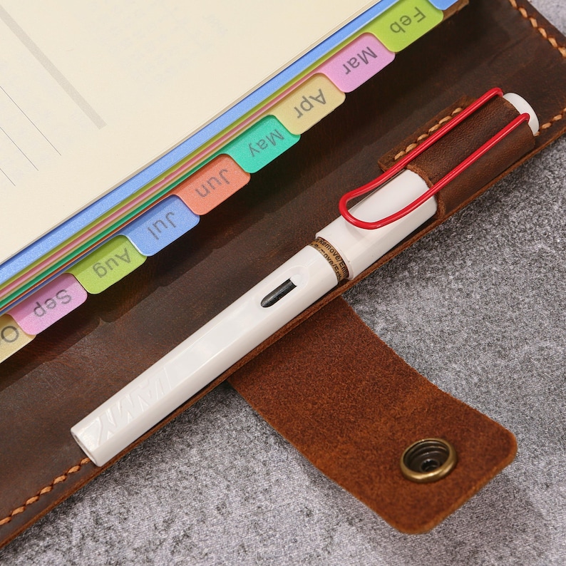 May include: A brown leather notebook cover with a snap closure and a pen loop holding a white fountain pen with a red wire clip. The notebook features colorful monthly index tabs labeled Feb, Mar, Apr, May, Jun, Jul, Aug, and Sep along the edge.