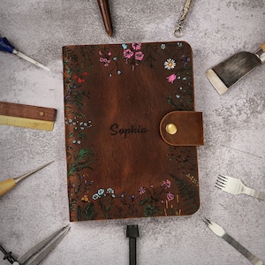 May include: A brown leather journal featuring a hand-painted floral border with pink, blue, and green accents. The name Sophia is embossed in the center. A brown leather strap with a brass snap button secures the cover, surrounded by various tools.