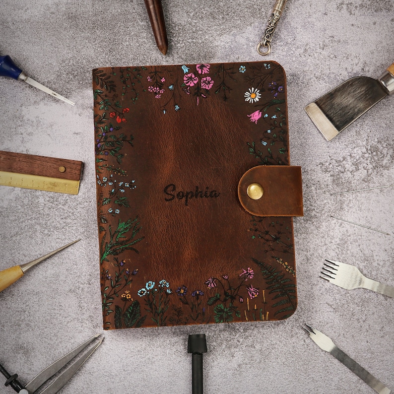 May include: A brown leather journal featuring a hand-painted floral border with pink, blue, and green accents. The name Sophia is embossed in the center. A brown leather strap with a brass snap button secures the cover, surrounded by various tools.