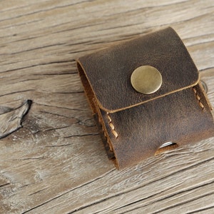 May include: A small, brown leather case with a brass button closure. The case has a distressed finish and visible stitching. The item is likely a small pouch or holder for accessories.