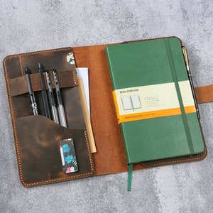 May include: A brown leather notebook cover with a green Moleskine notebook inside. The cover has a pocket for pens and a flap that closes with a button. The notebook has a ribbon bookmark and a pen loop.
