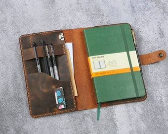 May include: A brown leather notebook cover with a green Moleskine notebook inside. The cover has a pocket for pens and a flap that closes with a button. The notebook has a ribbon bookmark and a pen loop.