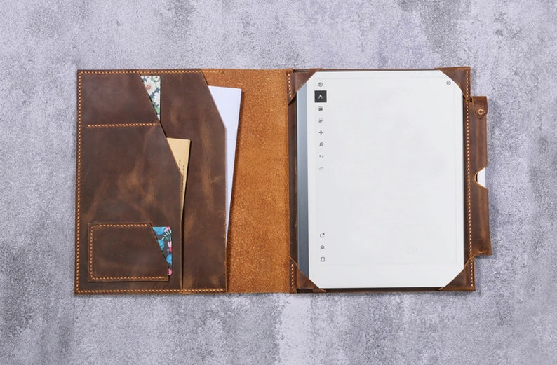 May include: A brown leather folio case featuring a left-side interior with multiple pockets for documents and cards, and a right-side sleeve holding a white digital tablet with a vertical menu bar and a side-mounted pen loop for a stylus.