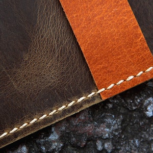 May include: Close-up of a brown leather surface with a contrasting orange leather strip sewn on top. The seam is stitched with white thread.