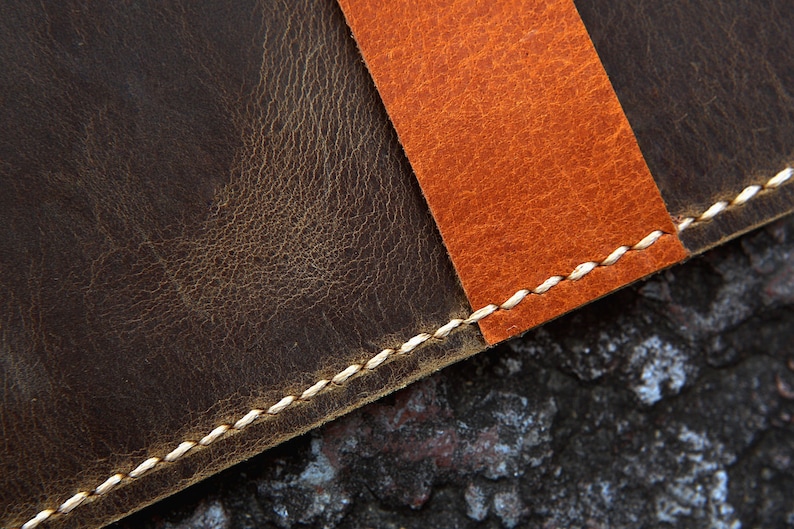 May include: Close-up of a brown leather surface with a contrasting orange leather strip sewn on top. The seam is stitched with white thread.