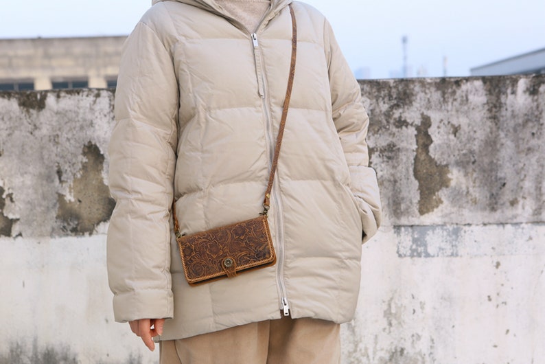 May include: A beige puffer jacket with a brown leather crossbody bag with an embossed floral design. The bag has a flap closure with a metal buckle.