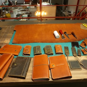 May include: A workbench with various leather goods, including wallets, keychains, and journals. There is a large piece of brown leather on the workbench, along with tools and supplies for leather crafting.