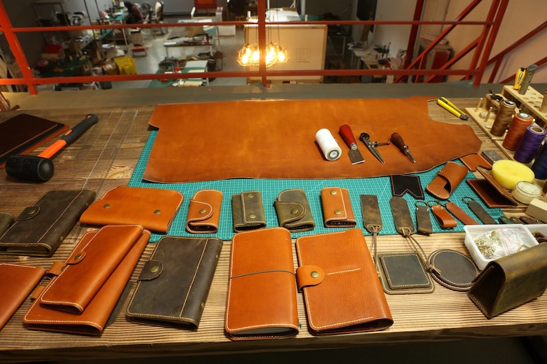 May include: A workbench with various leather goods, including wallets, keychains, and journals. There is a large piece of brown leather on the workbench, along with tools and supplies for leather crafting.