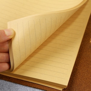 May include: A notebook with light brown, tan-colored paper pages featuring thin, dark horizontal lines. The pages are stacked and bound, with one corner of the top page lifted to reveal the lined texture and the consistent spacing of the paper.