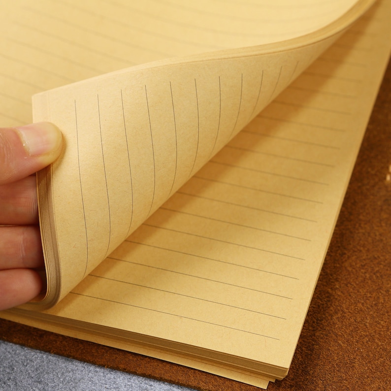 May include: A notebook with light brown, tan-colored paper pages featuring thin, dark horizontal lines. The pages are stacked and bound, with one corner of the top page lifted to reveal the lined texture and the consistent spacing of the paper.