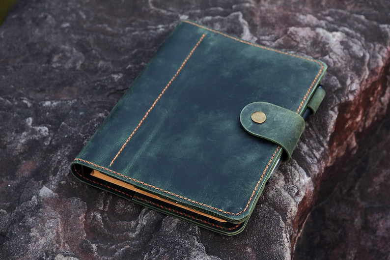 May include: A dark green leather bound journal with a brass button closure and orange stitching. The journal is lying on a textured gray rock.