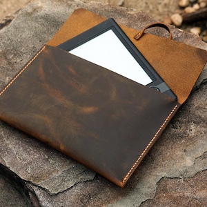 May include: A brown leather case with a flap and a stitched edge. The case is open and a black e-reader is inside.